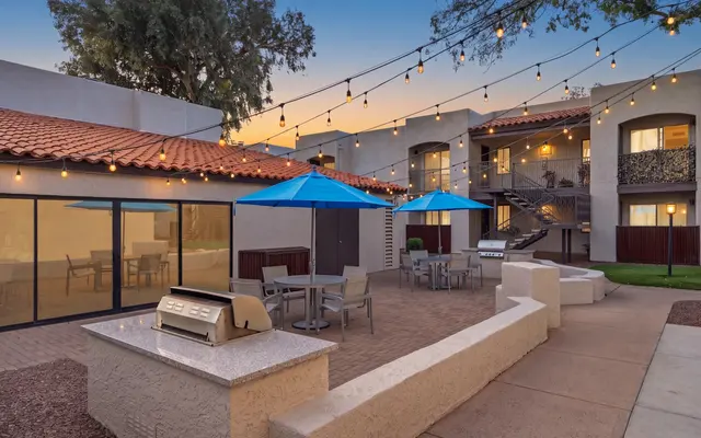 Outdoor communal area with a barbecue grill, seating under blue umbrellas, and string lights; an apartment building in the background.
