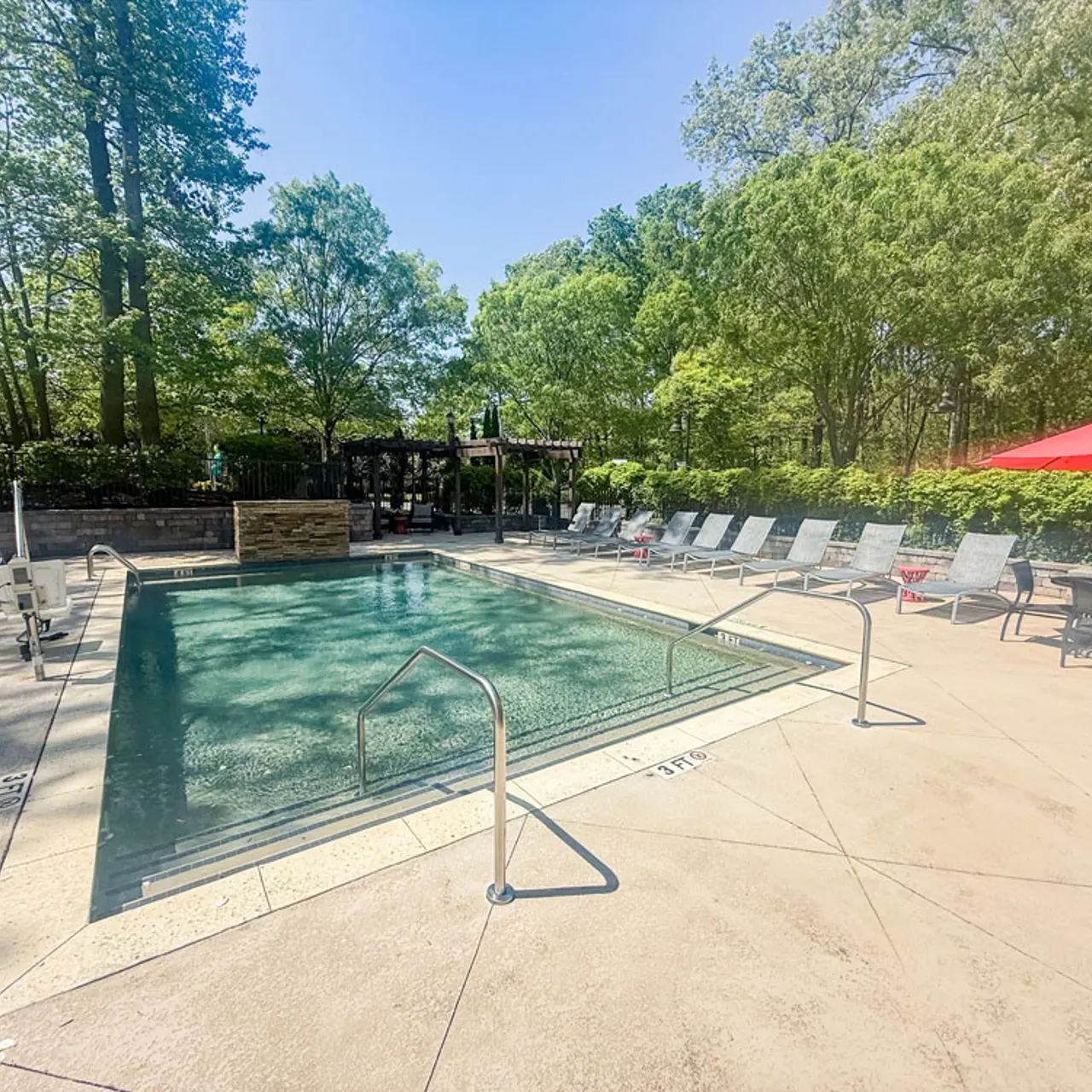 A tranquil swimming pool area surrounded by greenery, featuring a well-maintained pool with lounge chairs and an umbrella-covered seating area.