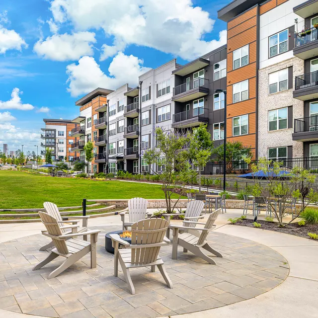 A modern apartment complex with multiple stories and balconies, surrounded by green grass and trees. In the foreground, there is a circular patio with several white Adirondack chairs arranged around a fire pit.