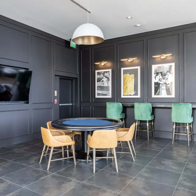 A modern interior space featuring a dark paneled wall, a round table with beige chairs, and green bar stools. There are framed artworks on the wall and a large TV mounted on the side.