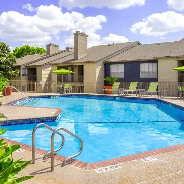 A clear swimming pool surrounded by lounge chairs and green umbrellas in a residential complex with blue skies and fluffy clouds in the background.