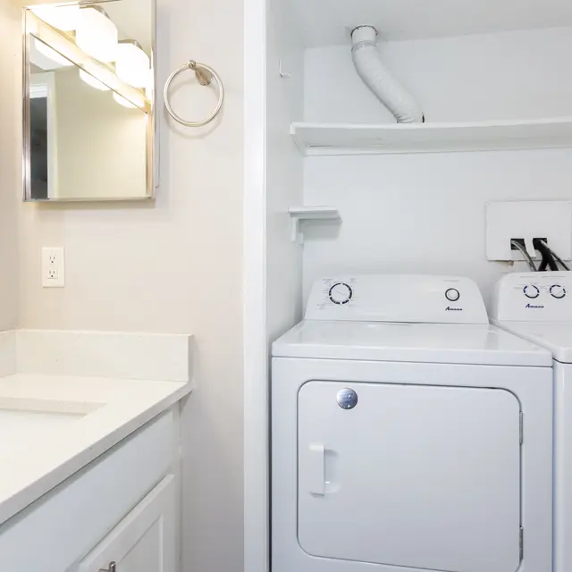 A clean and modern laundry room featuring a washer and dryer side by side, a white countertop, and a set of mirrors above a sink.