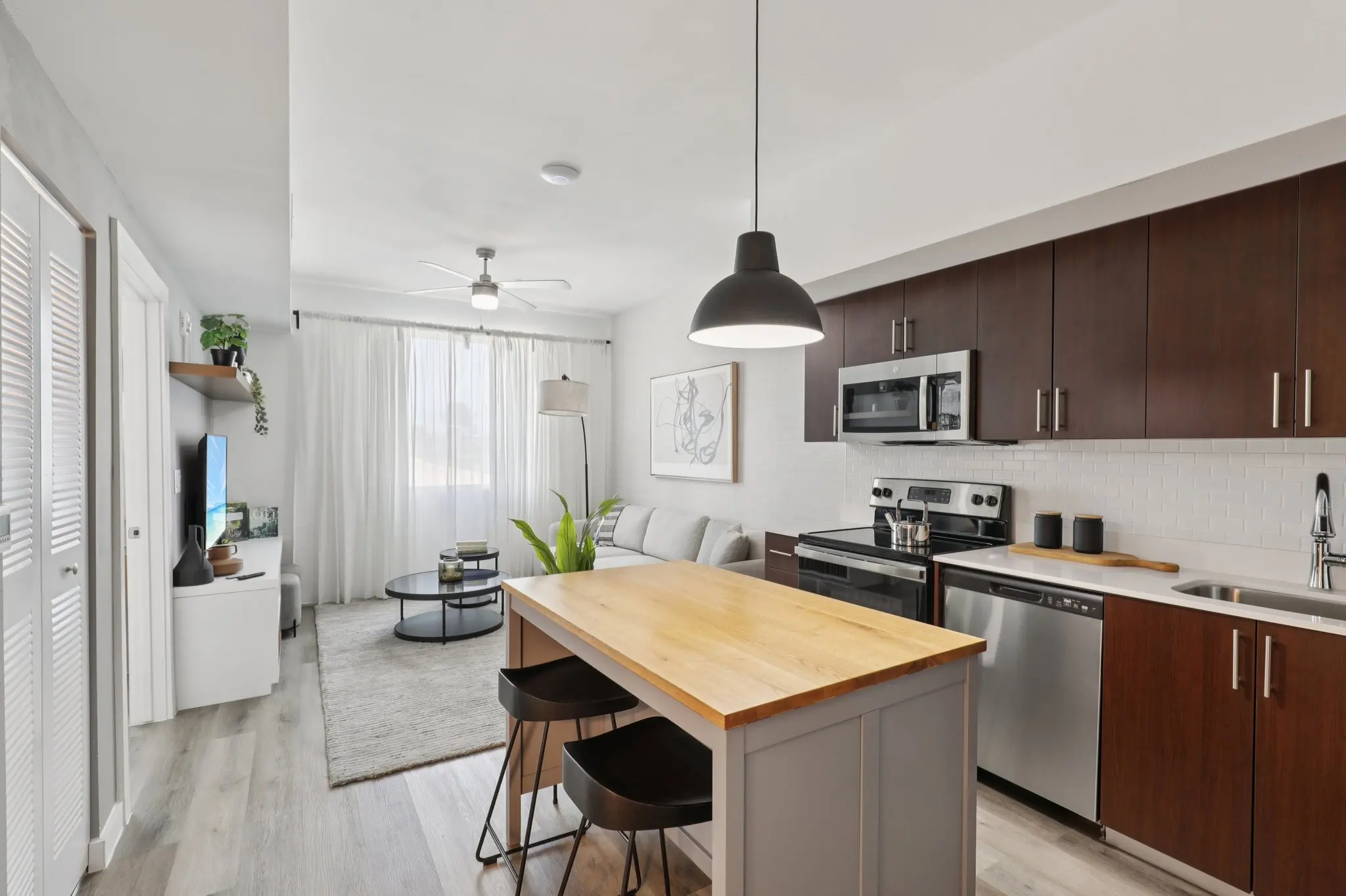 A well-lit modern apartment featuring a kitchen with dark cabinets, stainless steel appliances, and a wooden dining island with bar stools. The living area has a grey carpet, a cozy couch with a throw pillow, and a coffee table. Natural light comes through sheer curtains, enhancing the fresh decor.