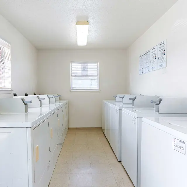 A clean and bright laundry room featuring several white washing machines and dryers arranged in a row. Natural light comes through the windows, illuminating the space which has beige tiled flooring and simple wall decorations with instructions or notices.