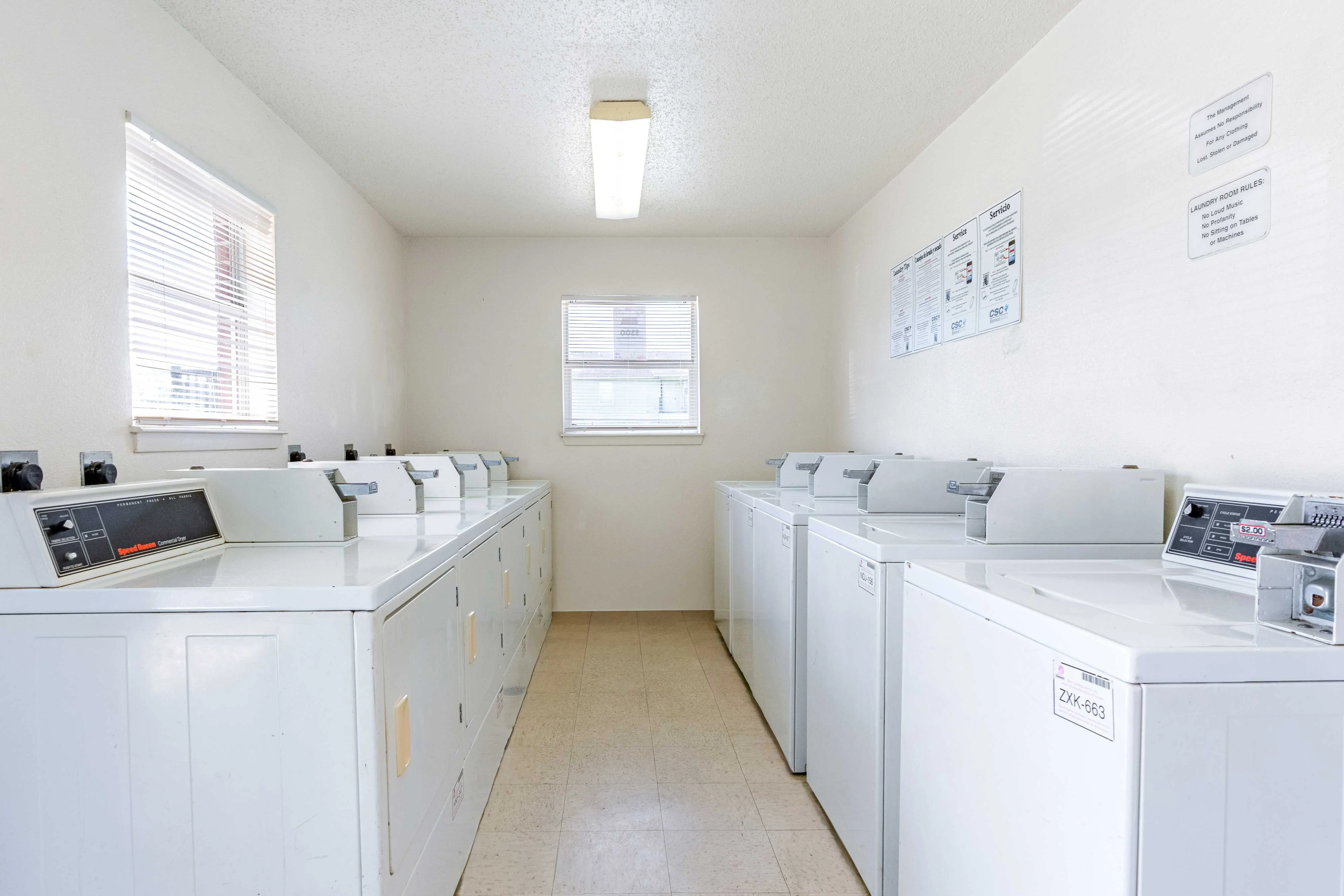 A clean and bright laundry room featuring several white washing machines and dryers arranged in a row. Natural light comes through the windows, illuminating the space which has beige tiled flooring and simple wall decorations with instructions or notices.