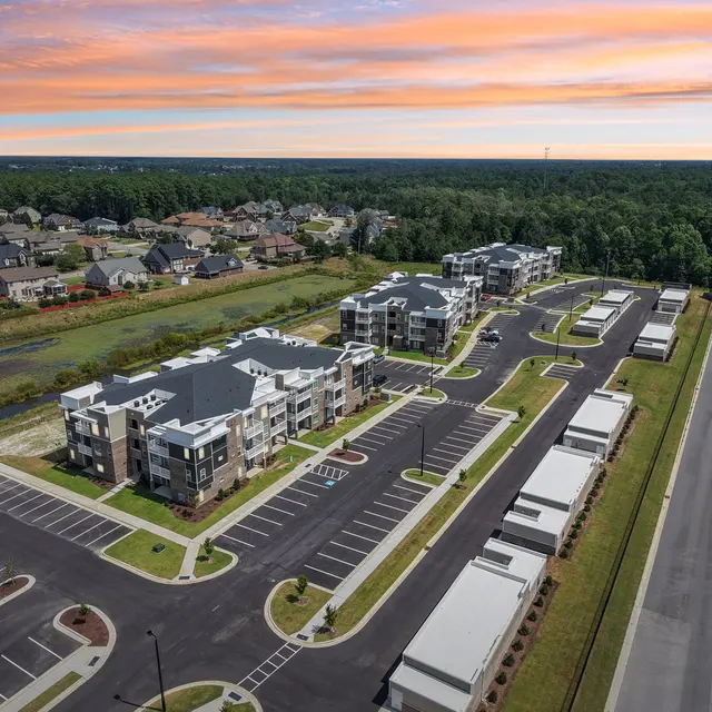 Aerial view of a modern apartment complex with multiple buildings and ample parking space, surrounded by greenery and residential areas under a colorful sunset sky.