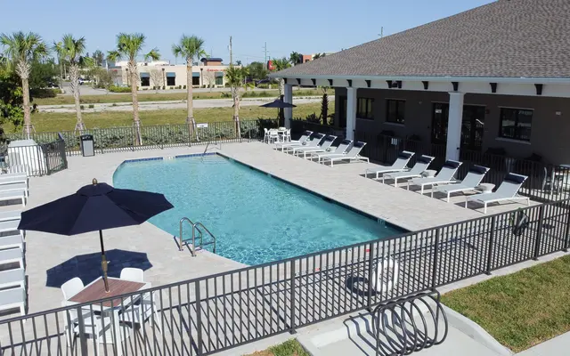 A swimming pool area featuring a clear blue pool surrounded by lounge chairs and an umbrella. There is a building in the background with large windows, and palm trees are visible around the area, under a clear blue sky.
