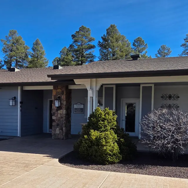 A modern house exterior with blue siding and stone accents. It features a well-kept garden with shrubs and a pathway leading to the entrance. Pine trees are visible in the background against a clear blue sky.