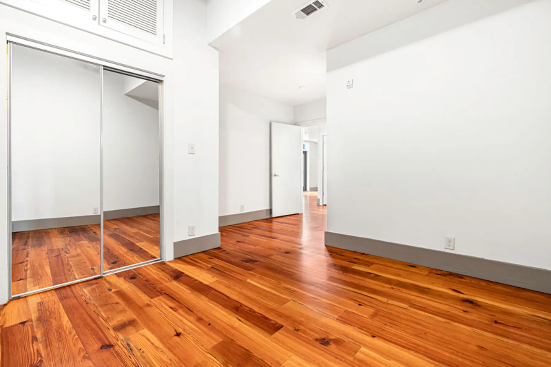 A spacious room featuring wooden flooring and bright white walls, with a large mirrored closet and a doorway leading to another room.