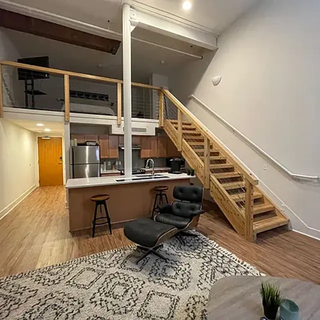 A stylish modern loft apartment featuring a wooden staircase leading to a loft area. The living space has a comfortable black chair, a rug, and a kitchenette with stainless steel appliances. Natural light fills the room from large windows.