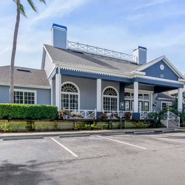 Exterior view of a blue building with decorative columns and a landscaped front area, featuring palm trees and parking spaces in front.