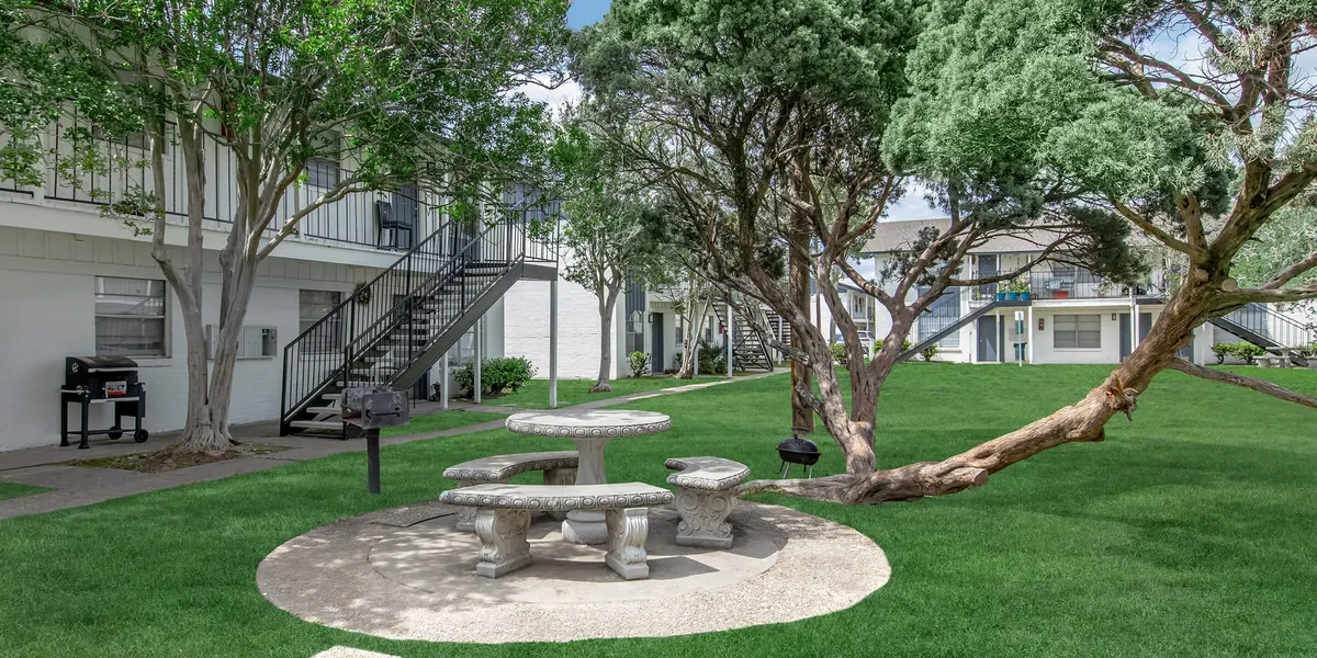 A landscaped outdoor area featuring a stone picnic table surrounded by grass and trees, with a staircase leading to an upper level in the background.