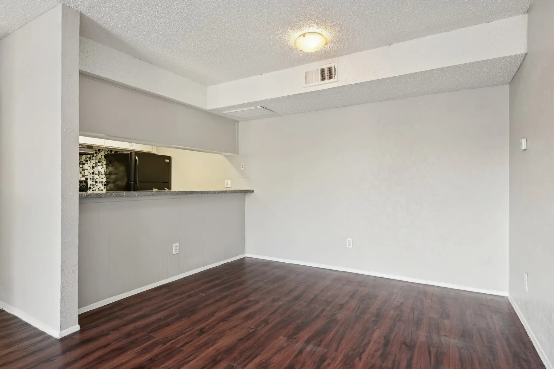 An empty living room with light beige walls and dark wooden flooring, featuring a small counter space leading to a kitchen area.