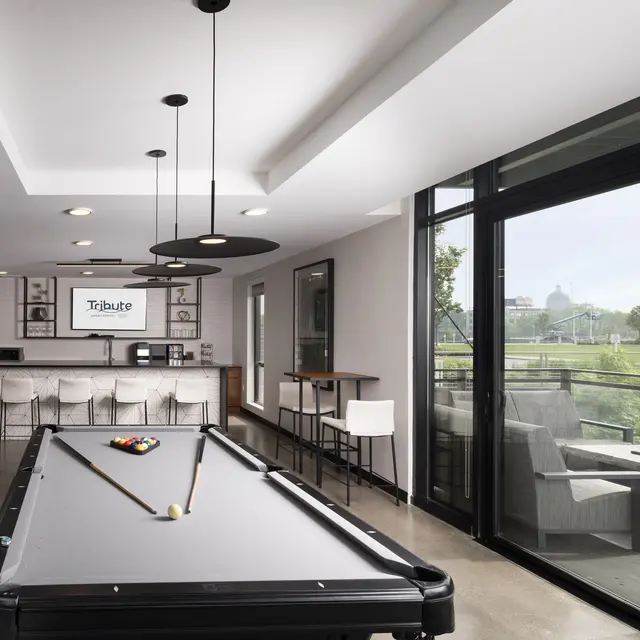 A modern lounge area featuring a billiards table and a large window with a view. The space includes a kitchen bar with stools and lounge seating.