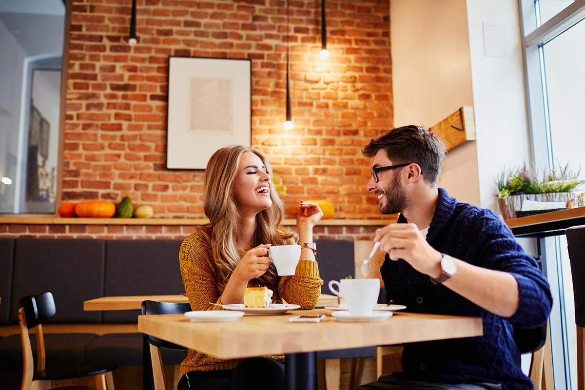 A couple sitting at a cafe table, smiling and enjoying coffee and dessert in a cozy environment with a brick wall.