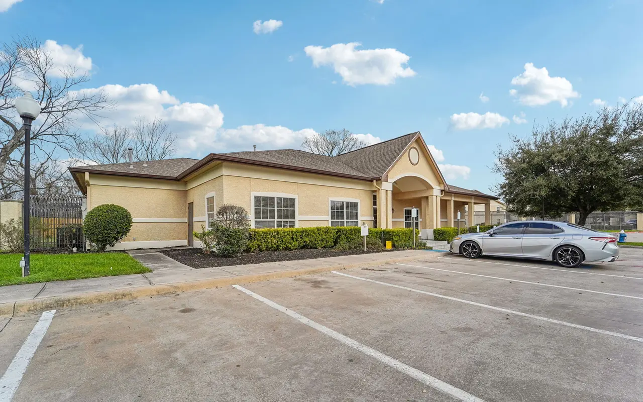 Exterior view of a commercial building with parking lot, featuring a beige facade and shrubbery, under a clear blue sky.