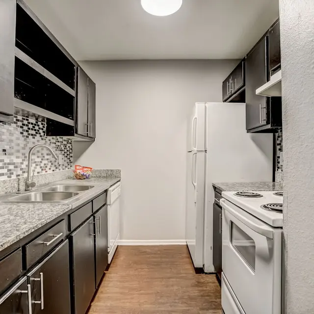A modern kitchen featuring dark cabinetry, granite countertops, and stainless steel appliances. The kitchen has a light wooden floor and textured backsplash.