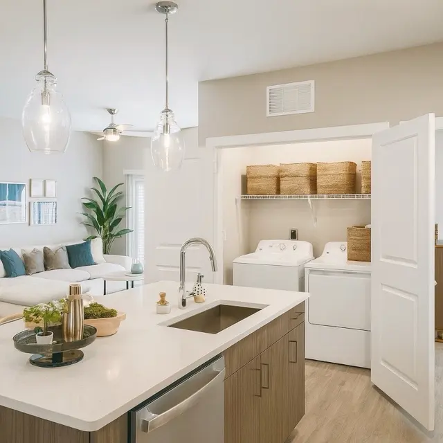 A modern kitchen featuring wooden cabinets, a white countertop, and a washer and dryer in a separate room. There are hanging light fixtures and a stylish living area visible in the background.
