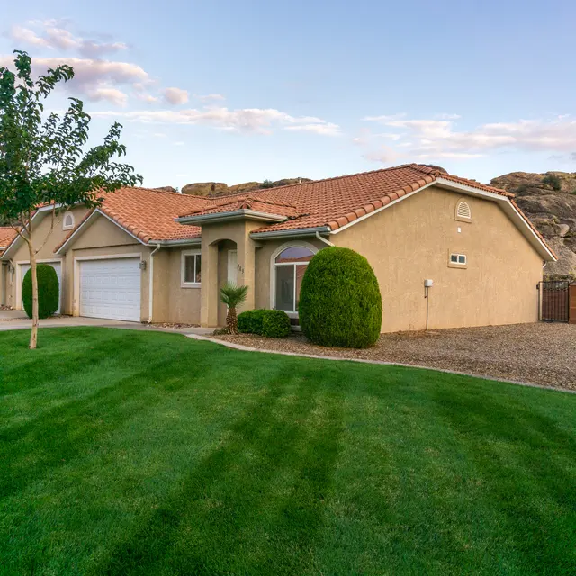 A single-story house with a red-tiled roof, surrounded by well-manicured green lawn and trees. In the background, there are large rocky formations under a partly cloudy sky.