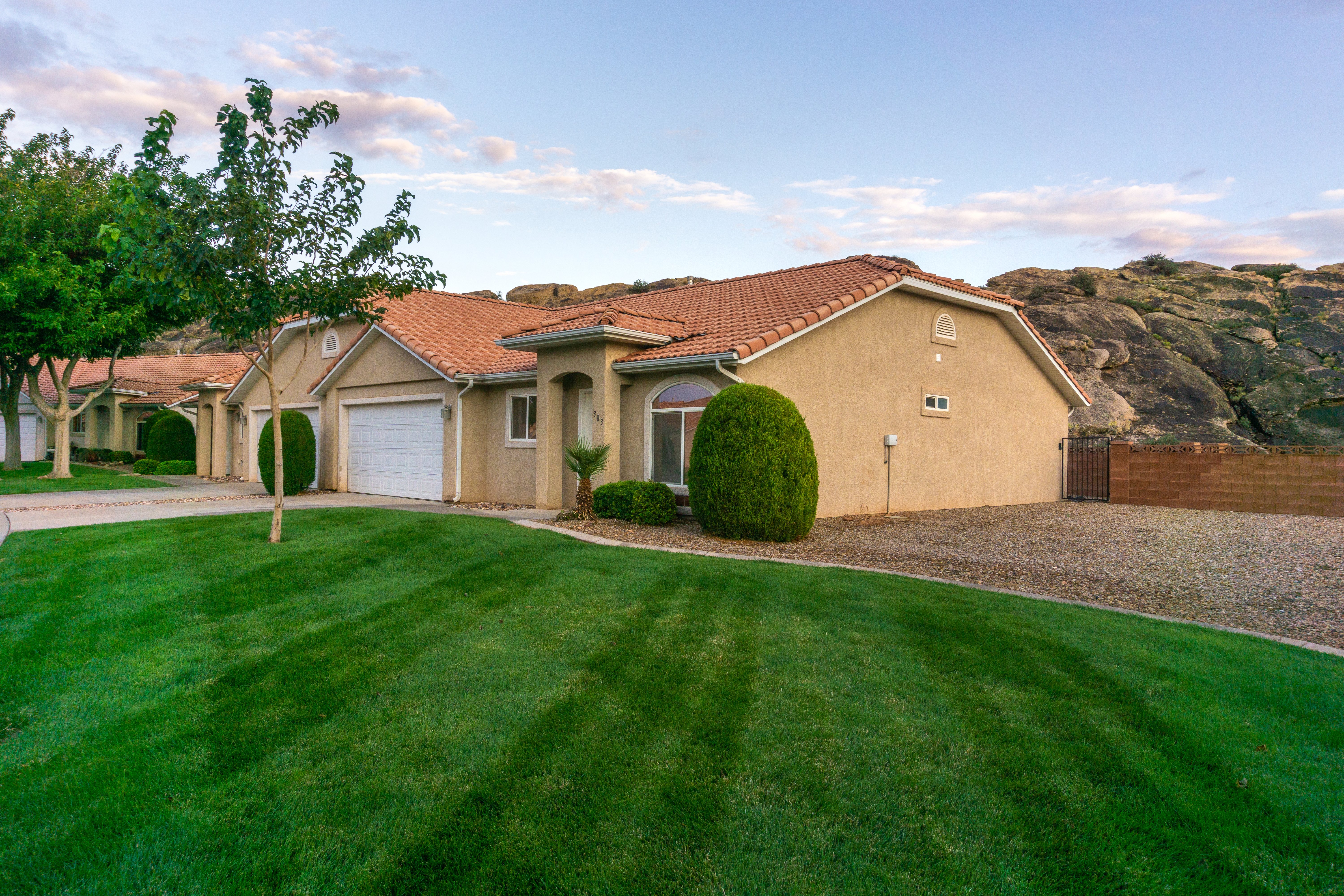 A single-story house with a red-tiled roof, surrounded by well-manicured green lawn and trees. In the background, there are large rocky formations under a partly cloudy sky.