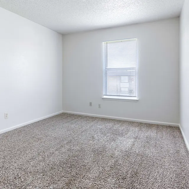 A vacant room with light gray walls and carpet, featuring a window on one side, empty with no furniture.