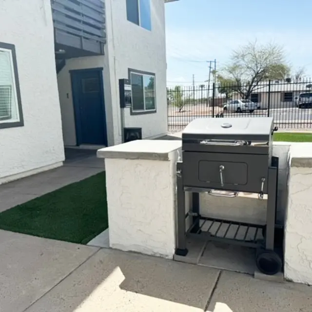 A modern outdoor grill located next to a white apartment building with a fenced yard and green turf nearby.