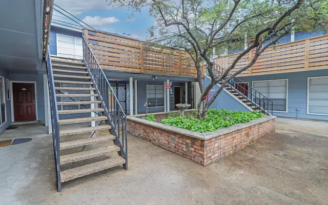A view of an apartment complex courtyard featuring a staircase, brick planters with greenery, and multiple doorways leading to units.