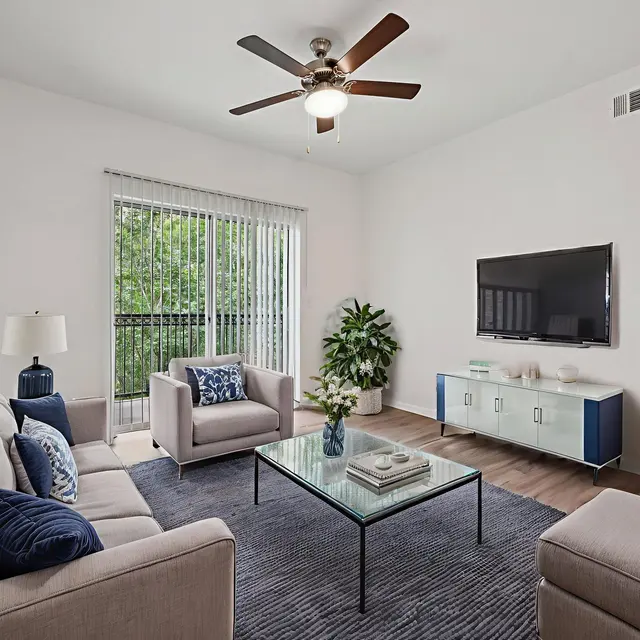 A modern living room featuring a gray sofa, beige chairs, a glass coffee table, and a wall-mounted television. A decorative plant adds a touch of greenery, with large windows letting in natural light.