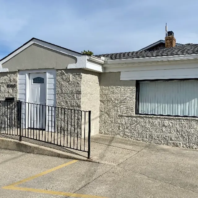 Exterior view of a single-story house with a light gray stone facade and white trim. The house has a ramp leading to the front door, which is white, and a large window with sheer curtains. There are two parking spaces visible in front.