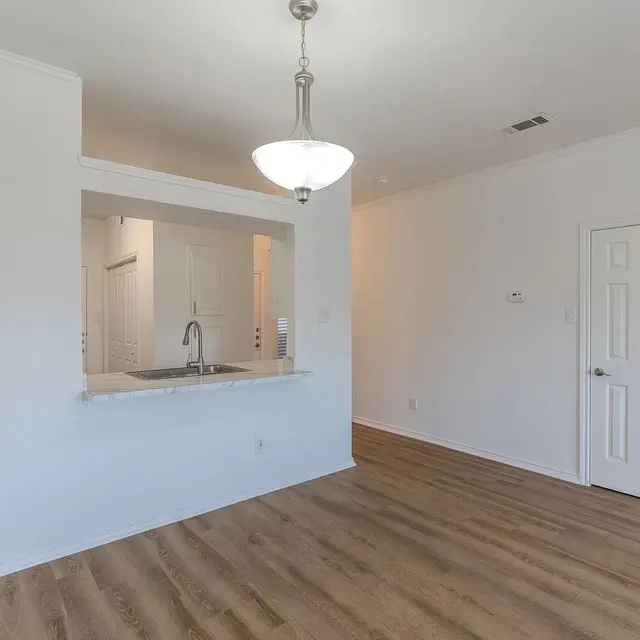 A modern living room featuring light-colored walls, wooden flooring, and a view of a kitchen area with a countertop. A pendant light hangs from the ceiling, illuminating the space.