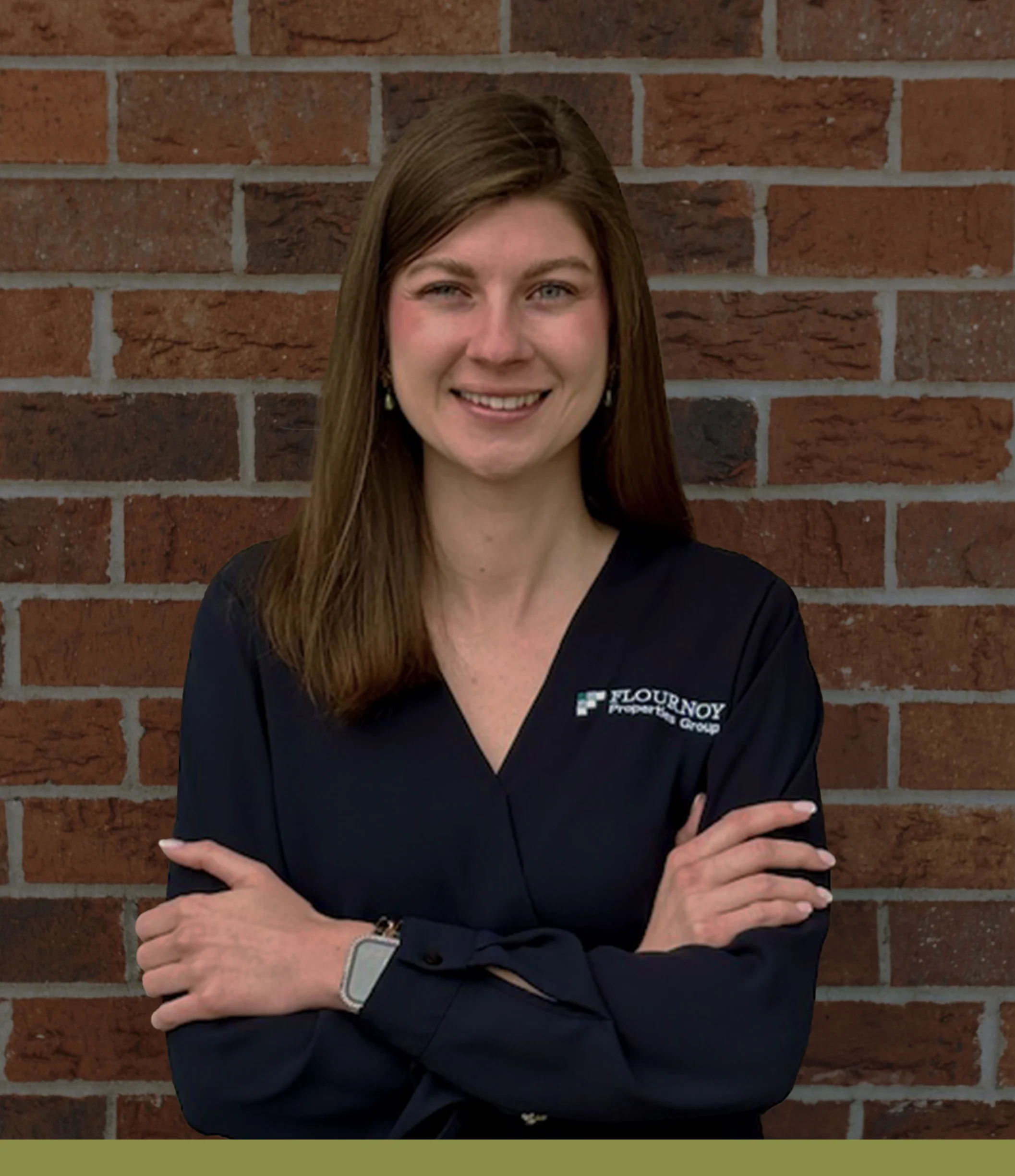 Professional Headshot A young woman with long brown hair smiles confidently with her arms crossed in front of a brick wall. She wears a dark-colored blouse with a logo on it.