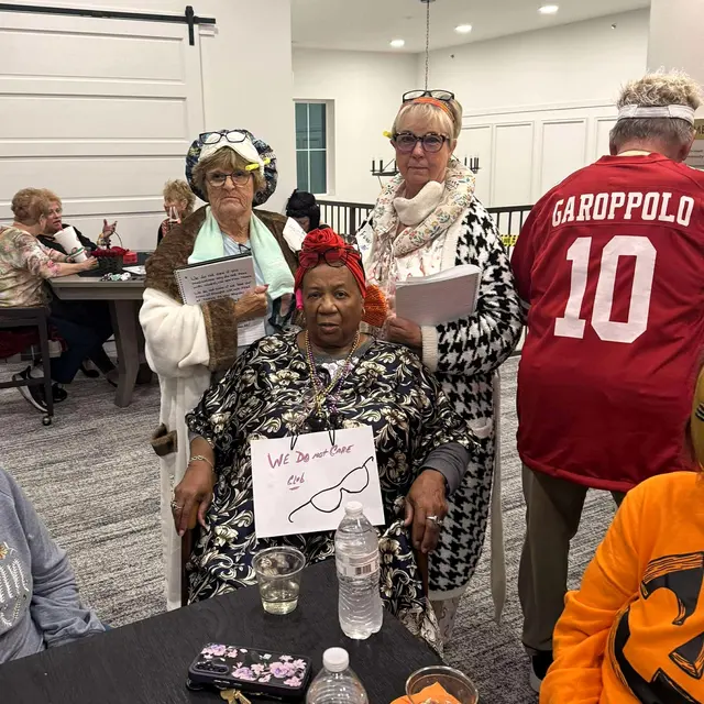 A group of elderly women and men dressed in themed outfits at a gathering, with a banner reading 'We Do Not Care Club' for Halloween.
