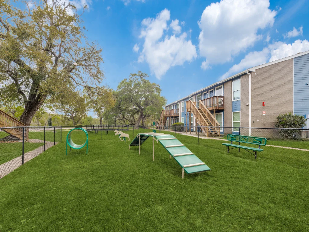 A spacious dog park with a green lawn, featuring an agility tunnel, a ramp, benches, and a multi-story apartment building in the background.
