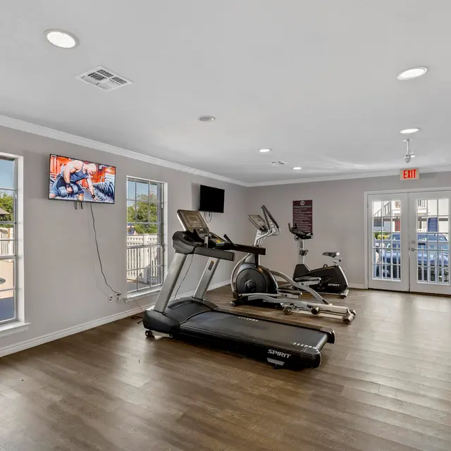 An interior view of a gym featuring two exercise machines (a treadmill and an elliptical), a television mounted on the wall, and large windows with natural light. The room has wooden flooring and is well-lit, with a door leading outside.