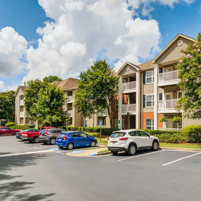 View of an apartment complex with parked cars and greenery under a bright blue sky with clouds