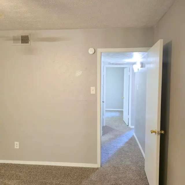 A view of a carpeted room with gray walls, featuring an open door that leads to a hallway. The door frame shows a glimpse of additional rooms in the background.