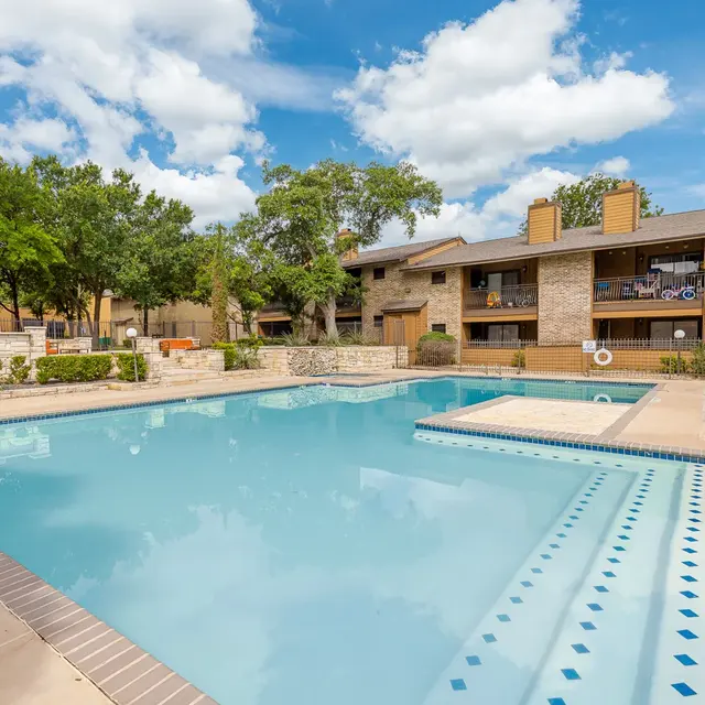 A clear swimming pool surrounded by a well-maintained apartment complex, with blue skies and fluffy clouds in the background.