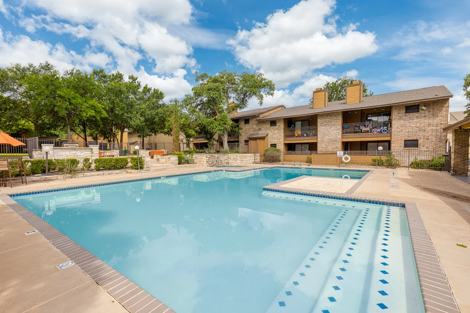 Apartment Complex Pool View A clear swimming pool surrounded by a well-maintained apartment complex, with blue skies and fluffy clouds in the background.