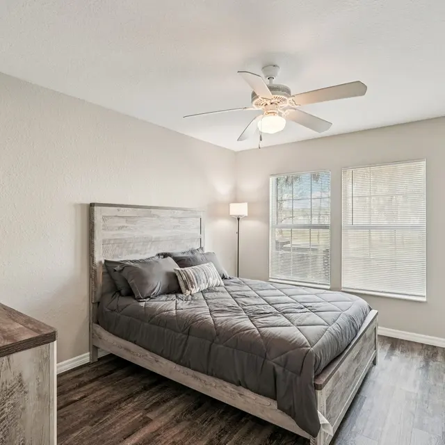 A modern bedroom with a bed, ceiling fan, and large windows. The bed has a gray comforter with a wooden headboard. A lamp stands beside the bed and there's a chest of drawers nearby.