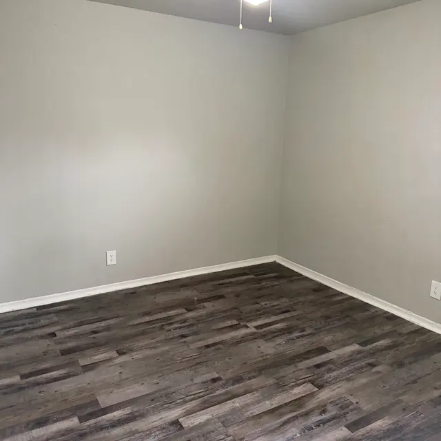 A spacious, empty room with light gray walls and a wooden floor. A ceiling fan is mounted in the center, providing a bright and airy feel. There are no furnishings or decorations in the room.