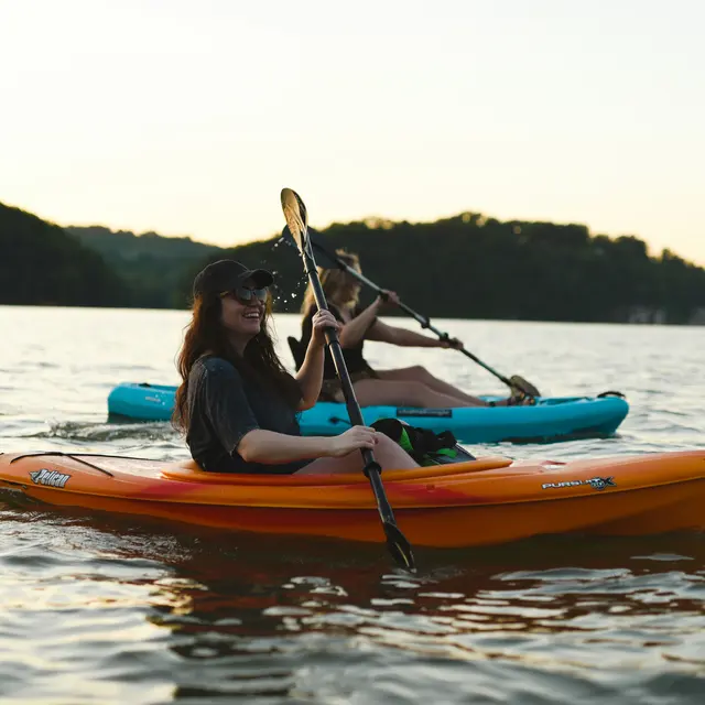 Milwaukee Kayak Company Two women kayaking on a calm lake during sunset. One is in an orange kayak, and the other is in a blue kayak, both smiling and enjoying their time on the water.