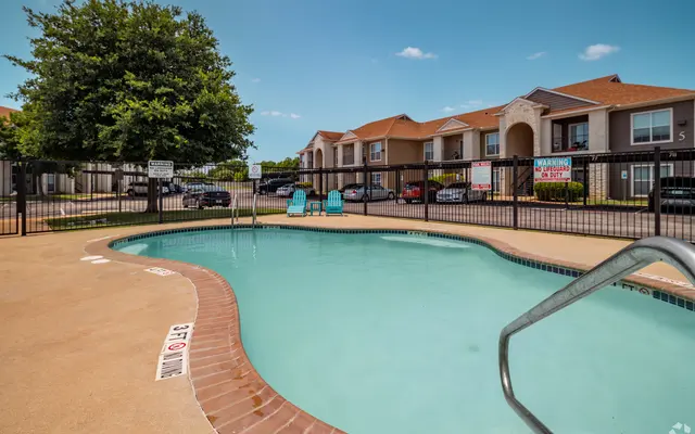 A small swimming pool area at an apartment complex, surrounded by a wooden fence and lounge chairs. In the background, several residential buildings are visible.