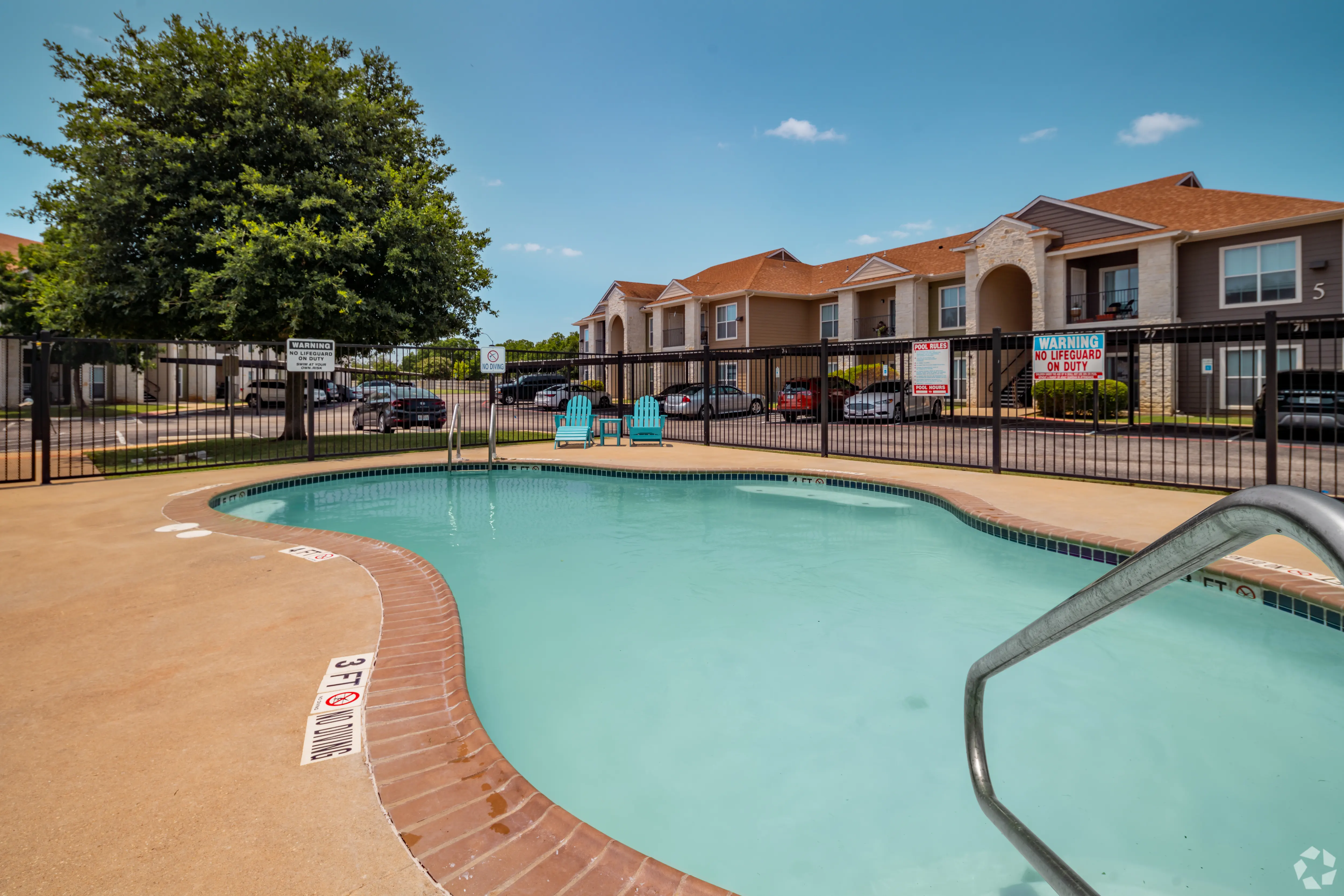 A small swimming pool area at an apartment complex, surrounded by a wooden fence and lounge chairs. In the background, several residential buildings are visible.