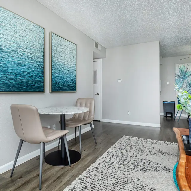 A modern dining area featuring a small round table with two beige chairs, surrounded by blue-toned abstract art on the walls. There is a light-colored rug on the floor, and a side table with decorative items. The space is bright and airy with natural light.