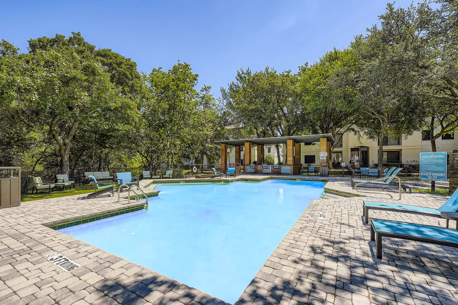 A clear blue swimming pool surrounded by lush green trees and lounge chairs on a sunny day. The pool area features a deck made of stone pavers and a shaded section with seating.