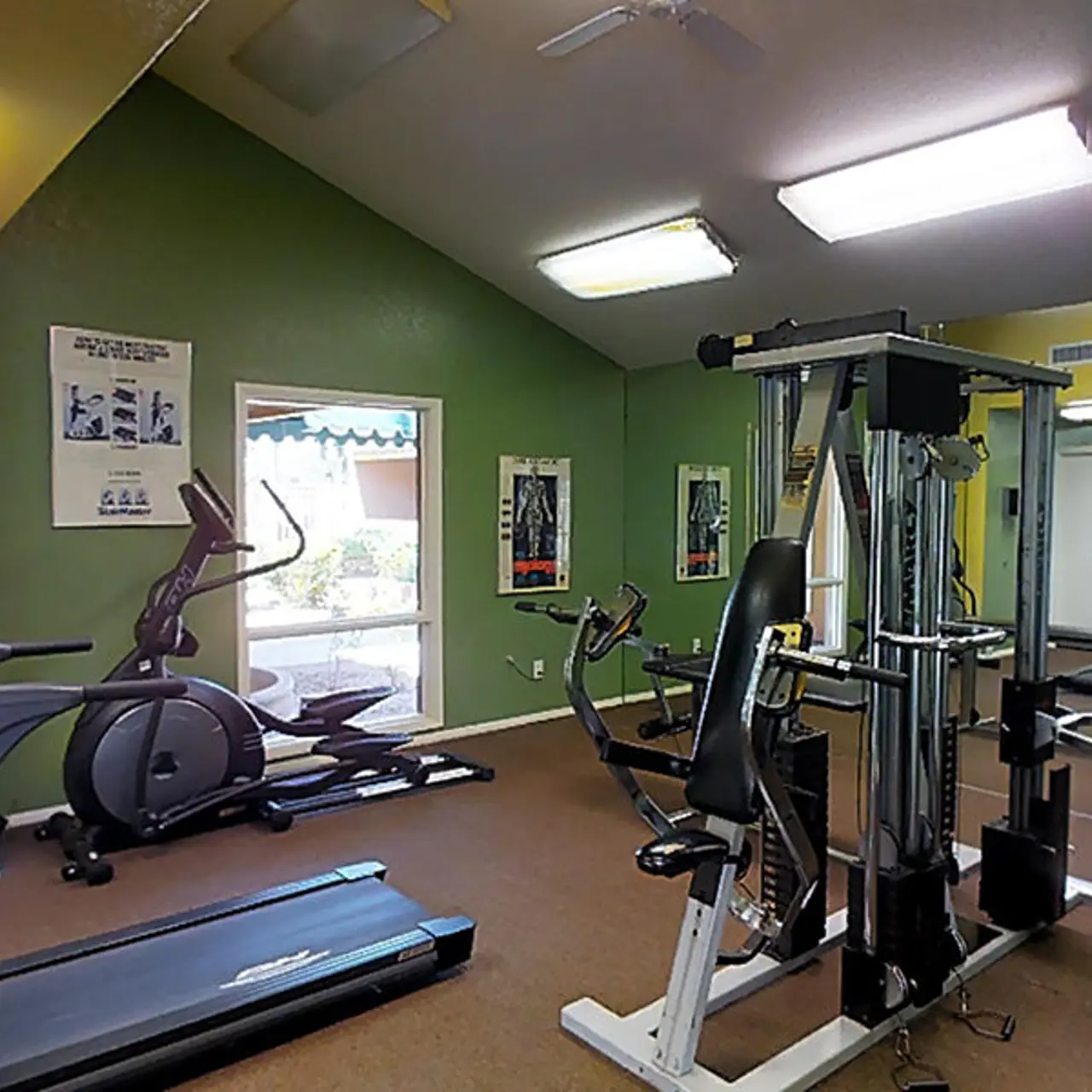 Interior view of a gym with various exercise equipment including a treadmill, elliptical machine, and weight training machine against green and yellow walls.
