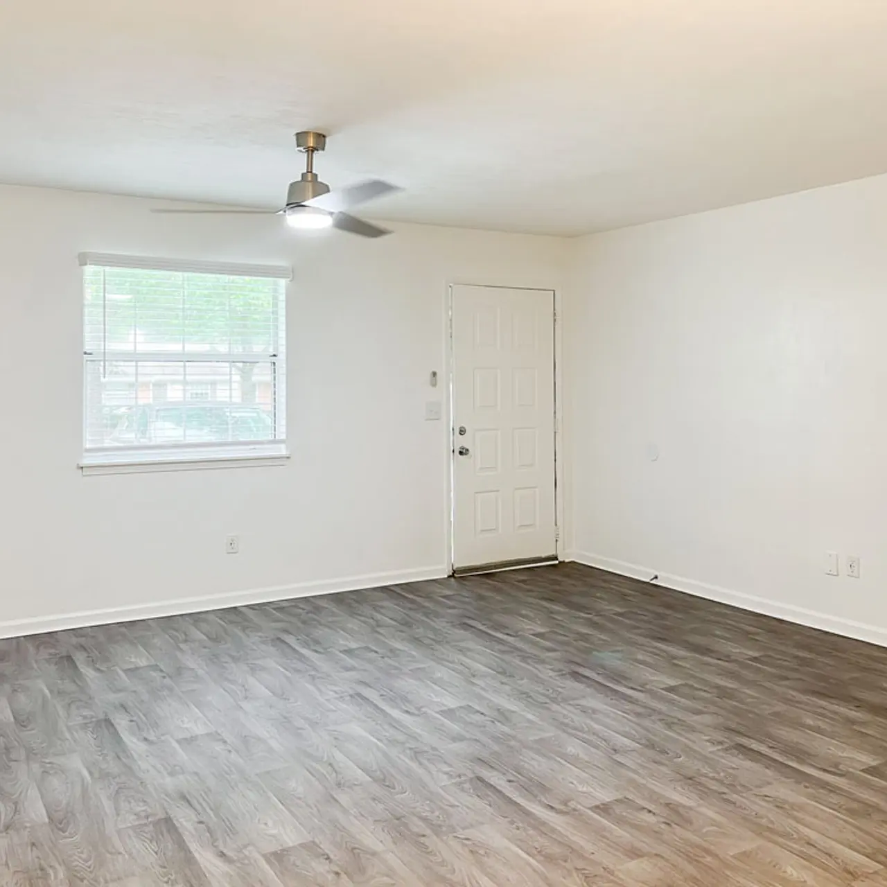 An empty living room featuring a ceiling fan, a white door, and a window with a view outside. The flooring is light-colored and resembles wood.