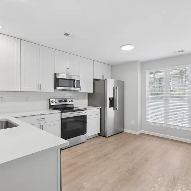 A modern kitchen with white cabinets, stainless steel appliances, and a large window with blinds that lets in natural light.