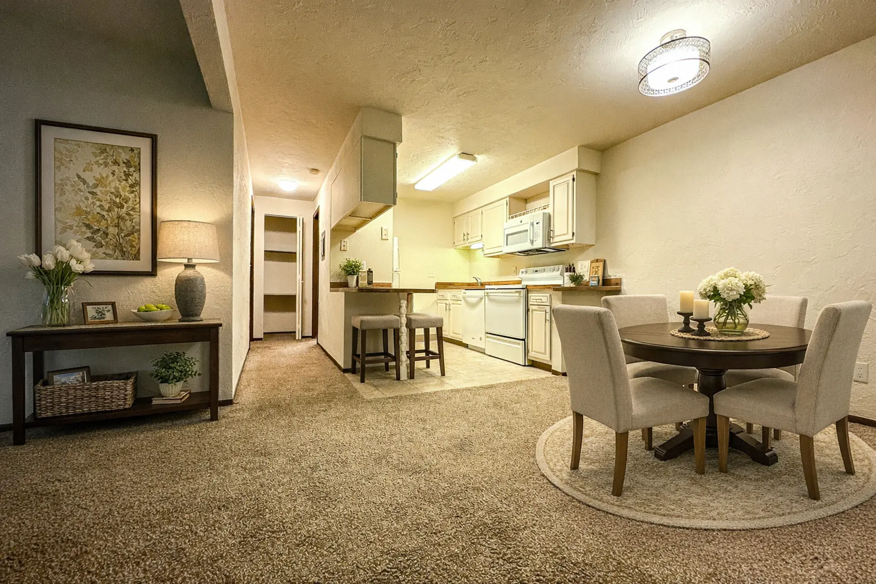 A spacious dining area connected to a modern kitchen. The room features a circular dining table with light gray chairs, a small kitchen island, and warm lighting. There is a couch near a decorative table with flowers and a framed artwork on the wall.