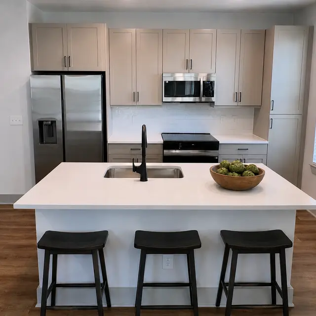 A modern kitchen with light-colored cabinets, stainless steel appliances, and a large island with bar stools. The island has a bowl of fruit on it.